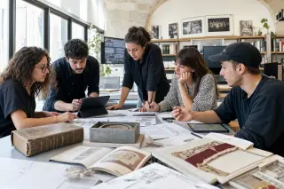 Creative professionals collaborating around a table in a modern coworking space with heritage details, exchanging ideas amid digital tools and historical artifacts under natural light.