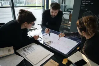 Professionals collaborating around an illuminated interactive table in a digital lab, engrossed in creative work with archival materials and digital maps