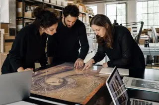 Three professionals collaborate intensely over a touchscreen table with a digital painting in a creative heritage lab, surrounded by archival tools and illuminated by soft natural light.