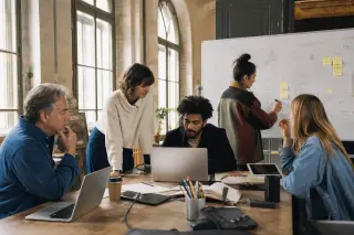 Creative professionals of different ages collaborating at a sunlit coworking table, interactively blending heritage items with digital technology in a lively brainstorming session.