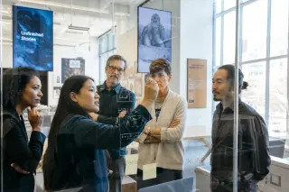 Museum and business professionals collaborating around a glass board in a bright, modern coworking space, with natural light and a creative, professional atmosphere.