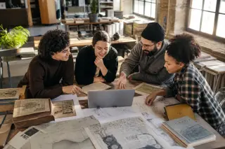 Group of four heritage professionals in a creative coworking space collaborating around a laptop and historical materials, engaged in discussion with natural light flooding the room.