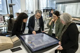 Heritage professionals collaborating around a touchscreen table in a digital lab, engaged in annotating scanned manuscripts with artifacts and tech equipment visible in a softly lit, modern space.